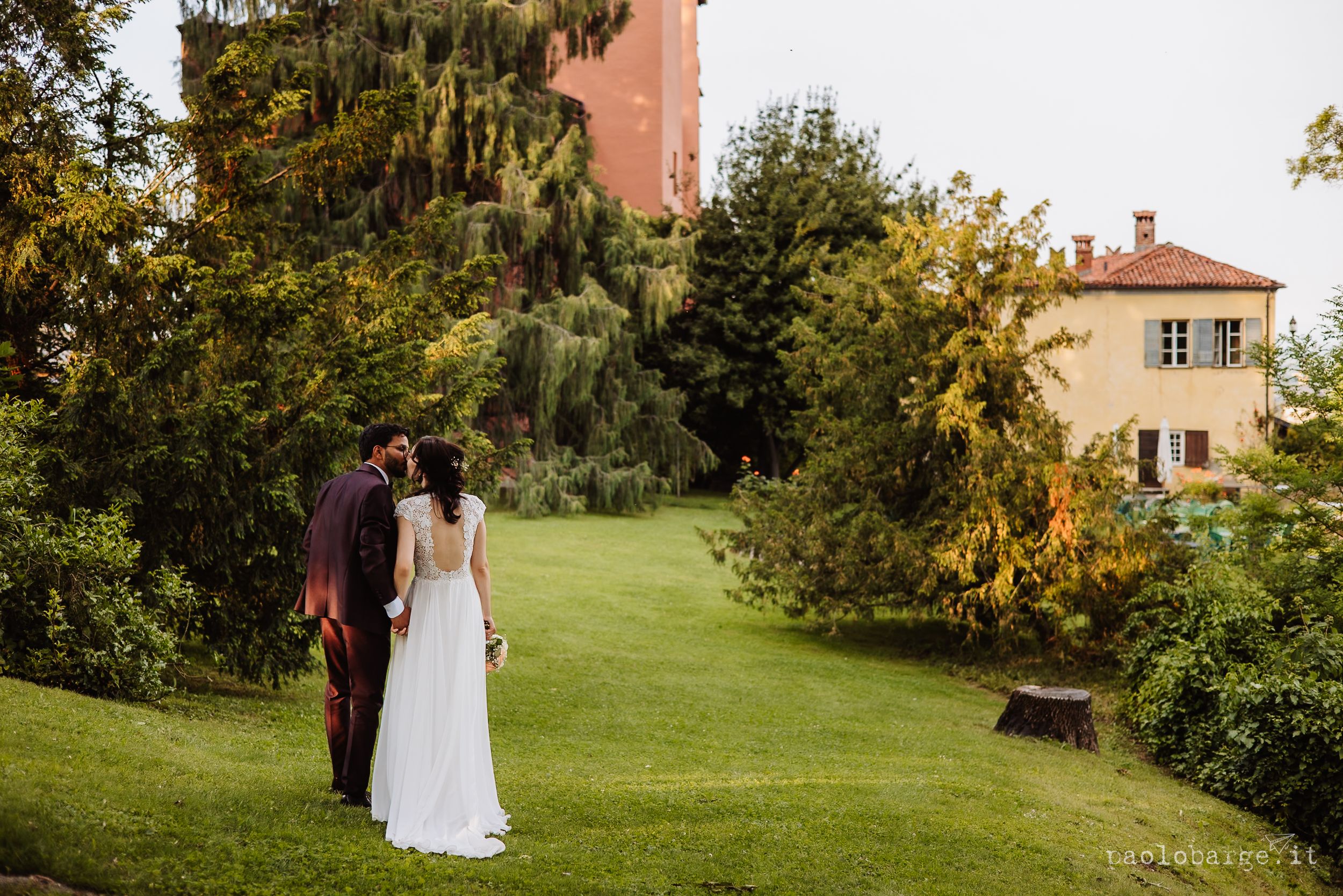 A couple in white dresses walks together across a lush garden lawn surrounded by trees and buildings.