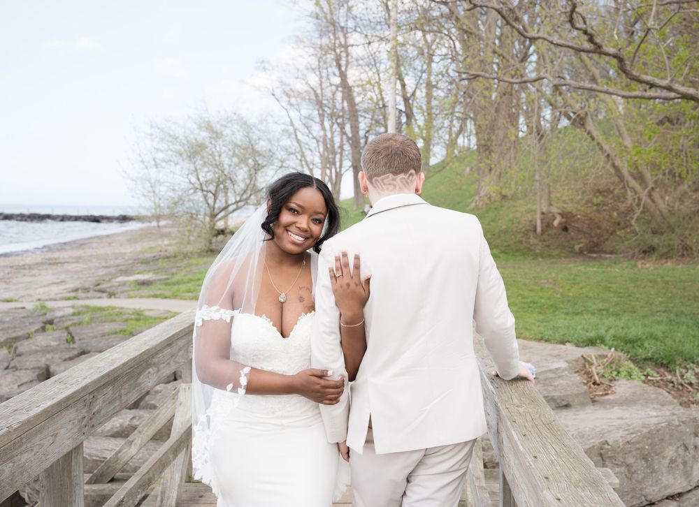 A smiling couple in wedding attire share a tender moment together on a waterfront path by bare trees in early spring.