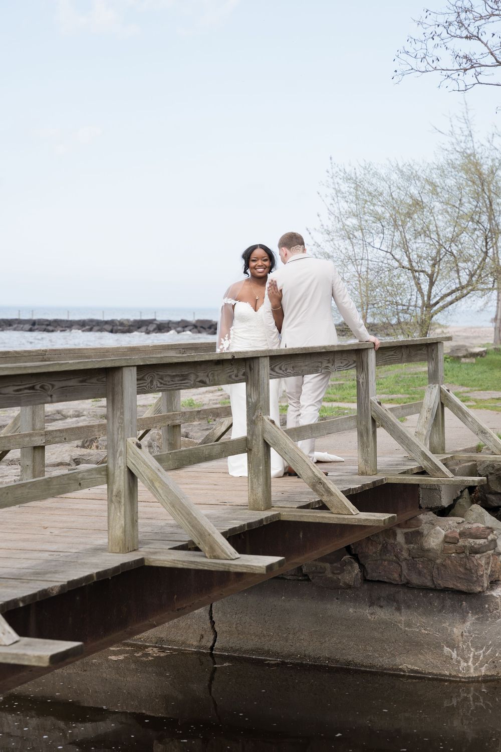 A couple in wedding attire stands together on a rustic wooden bridge overlooking a waterfront on their special day.