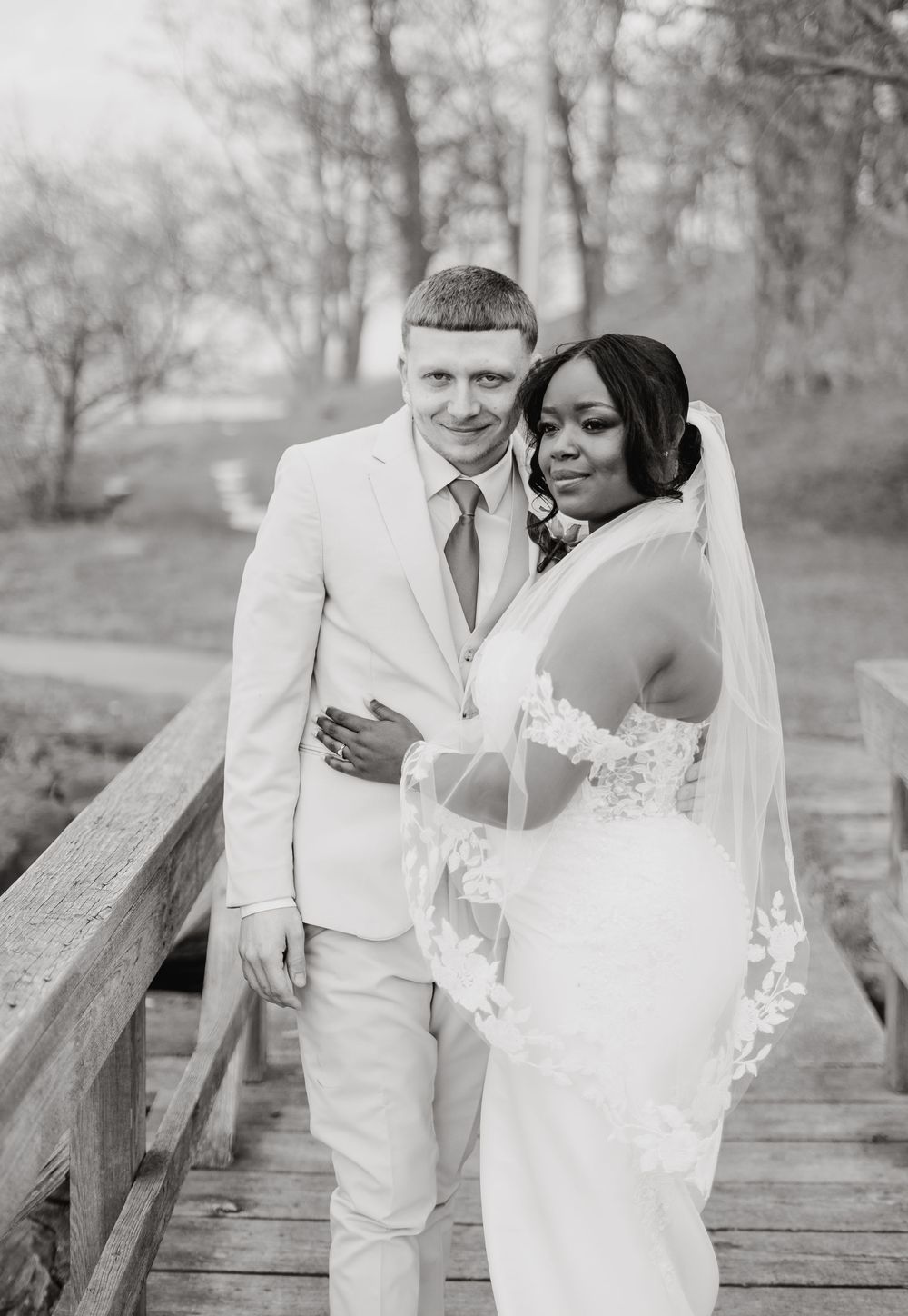 A happy couple pose together on a wooden bridge for their black and white wedding portrait in a wooded setting.