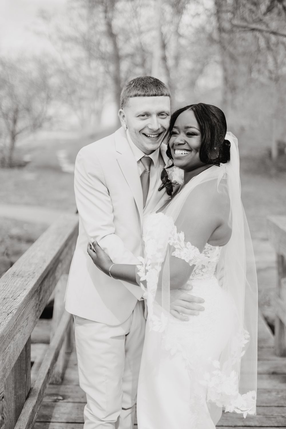 A joyful bride and groom embrace and laugh together on a wooden bridge in black and white photography.