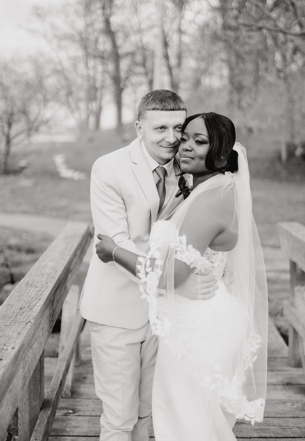 A black and white photo captures a romantic wedding moment of a couple embracing on a wooden bridge in nature.
