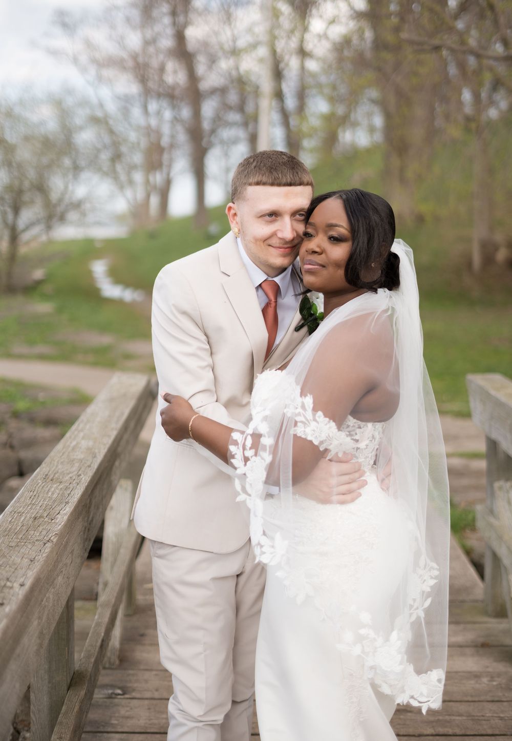 A romantic wedding photo of a couple in formal attire embracing on a wooden bridge surrounded by trees.