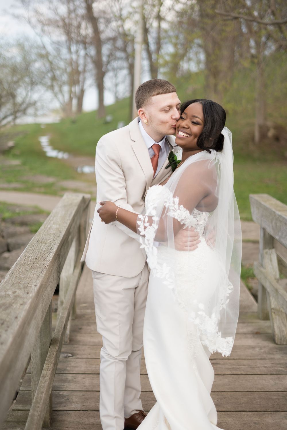 A romantic wedding couple embracing on a rustic wooden bridge surrounded by spring trees and greenery.