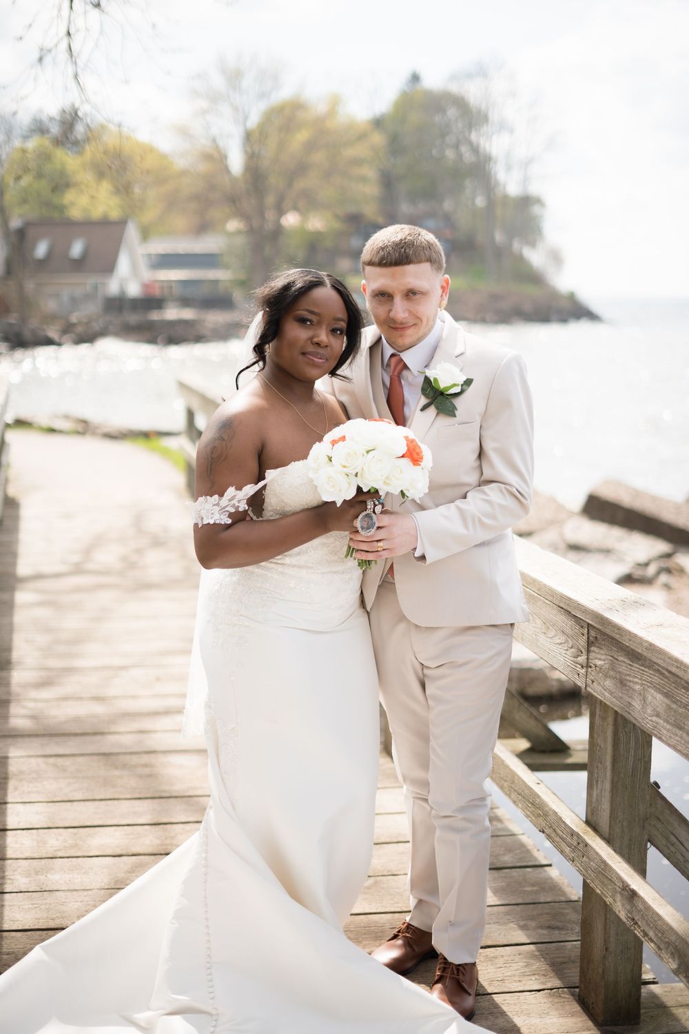 A couple in wedding attire sharing a tender moment on a coastal boardwalk with water and trees in the background.