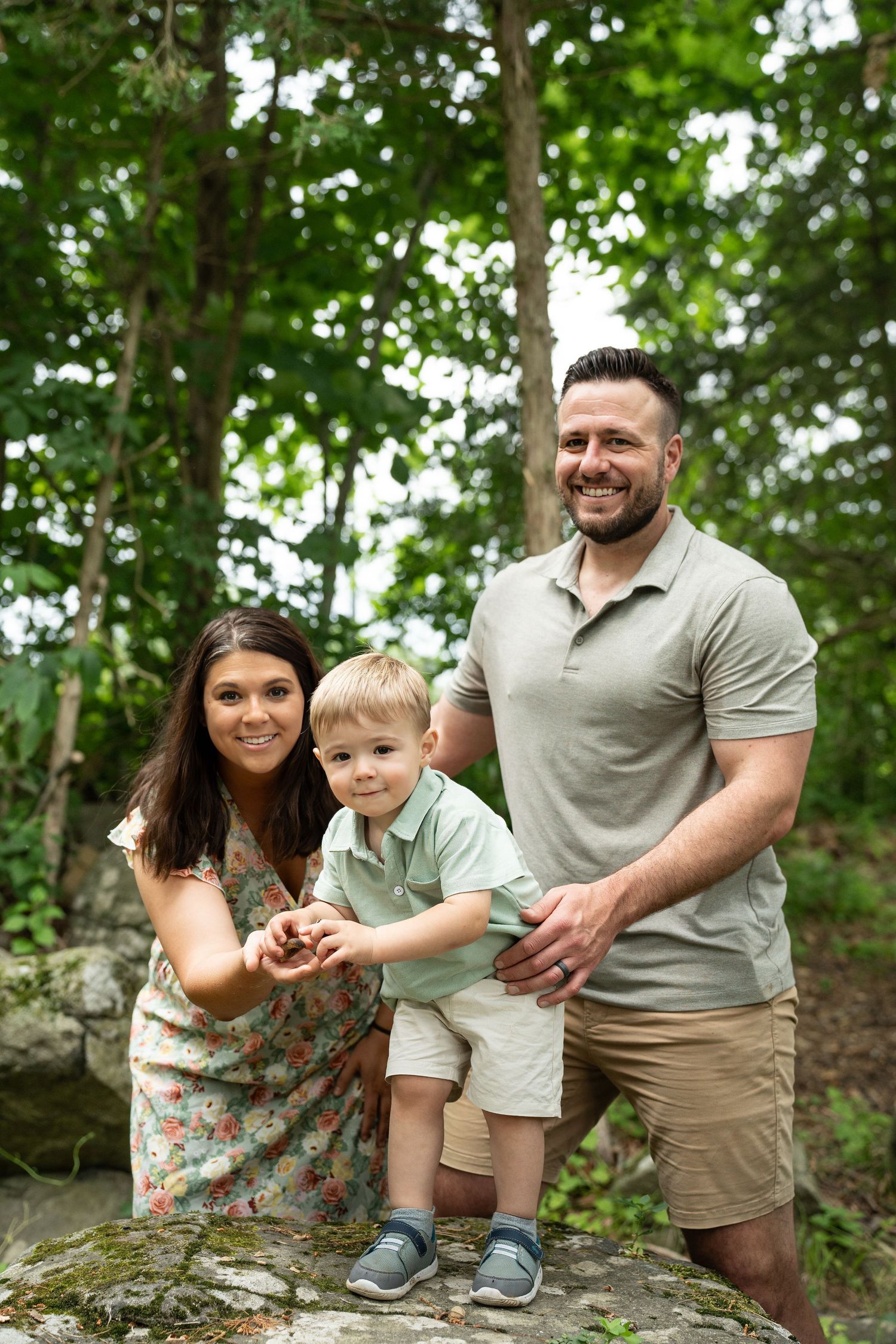 A family of three poses together on a rock outdoors in a wooded setting wearing casual summer attire. Child is playing with rocks.