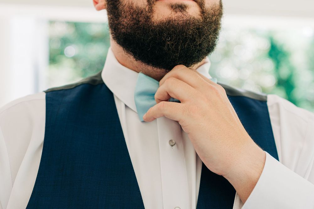 A close-up view of someone adjusting their bowtie while wearing a navy vest and white dress shirt.