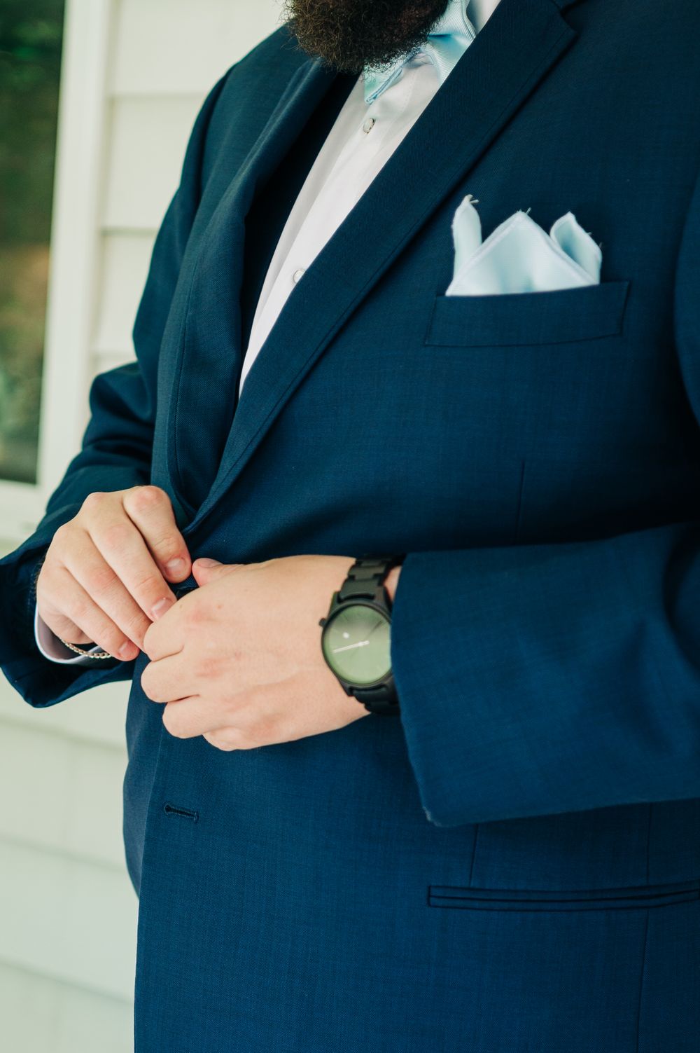 Close-up of navy blue suit jacket with white pocket square and wristwatch detail on formal attire.