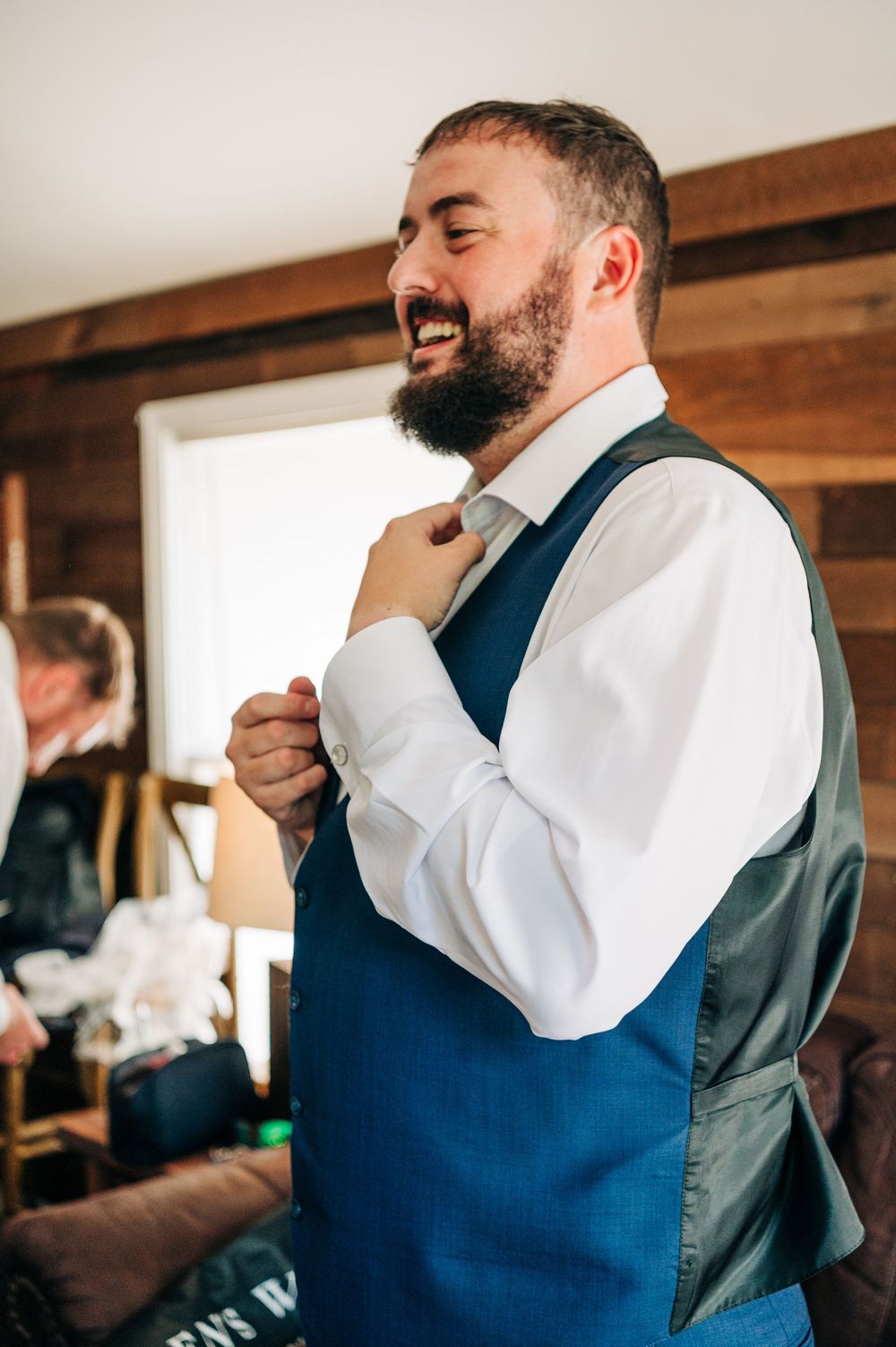 A person in a blue vest and white dress shirt laughs while adjusting his tie during what appears to be formal preparation.
