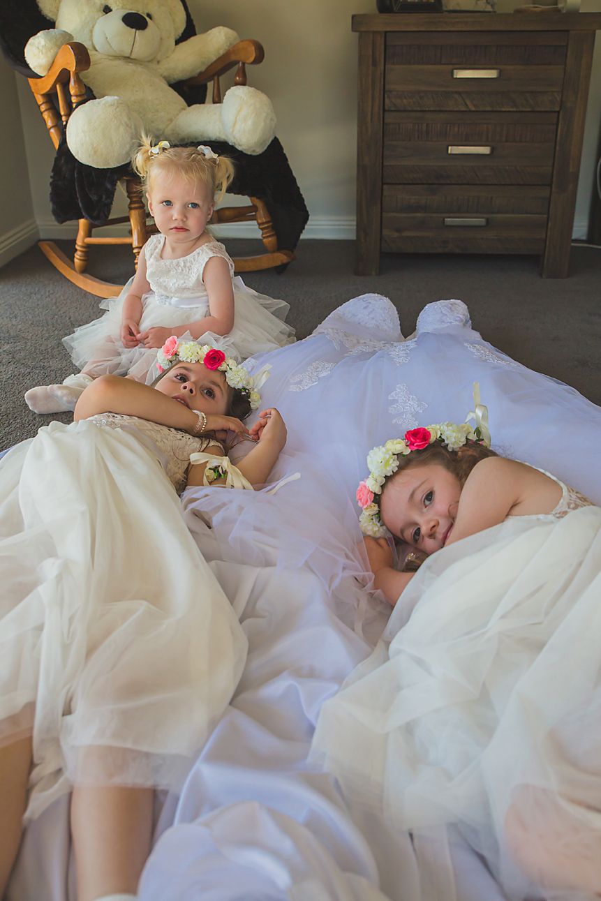 Two individuals wearing white dresses laying on tulle fabric with floral crowns near a wooden dresser and teddy bear.