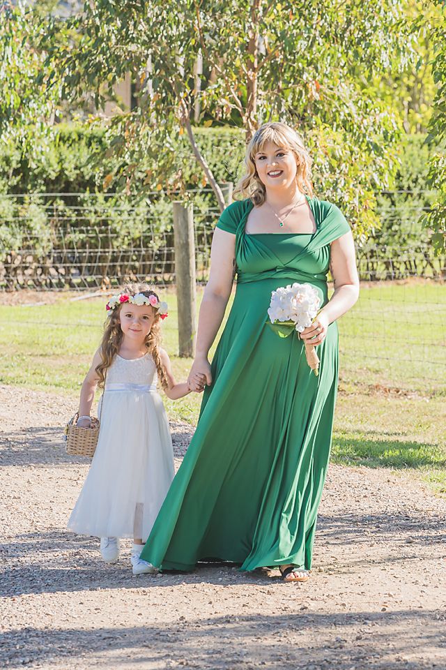 A pair in white and emerald formal dresses standing together on a garden path with floral accessories.