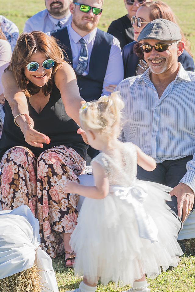 A flower girl in a white dress plays and twirls on the grass while wedding guests in sunglasses look on and smile.