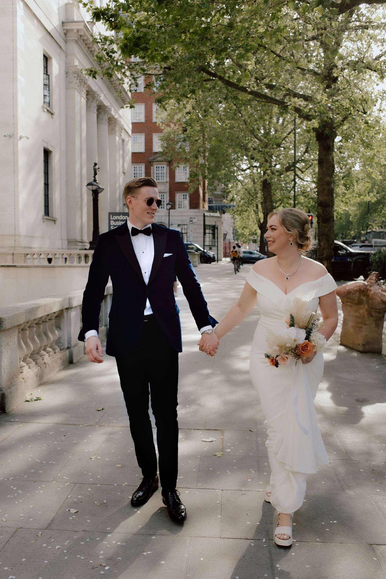 A couple walks down white marble stairs on their wedding day, with the bride in white holding a bouquet and groom in navy tuxedo.