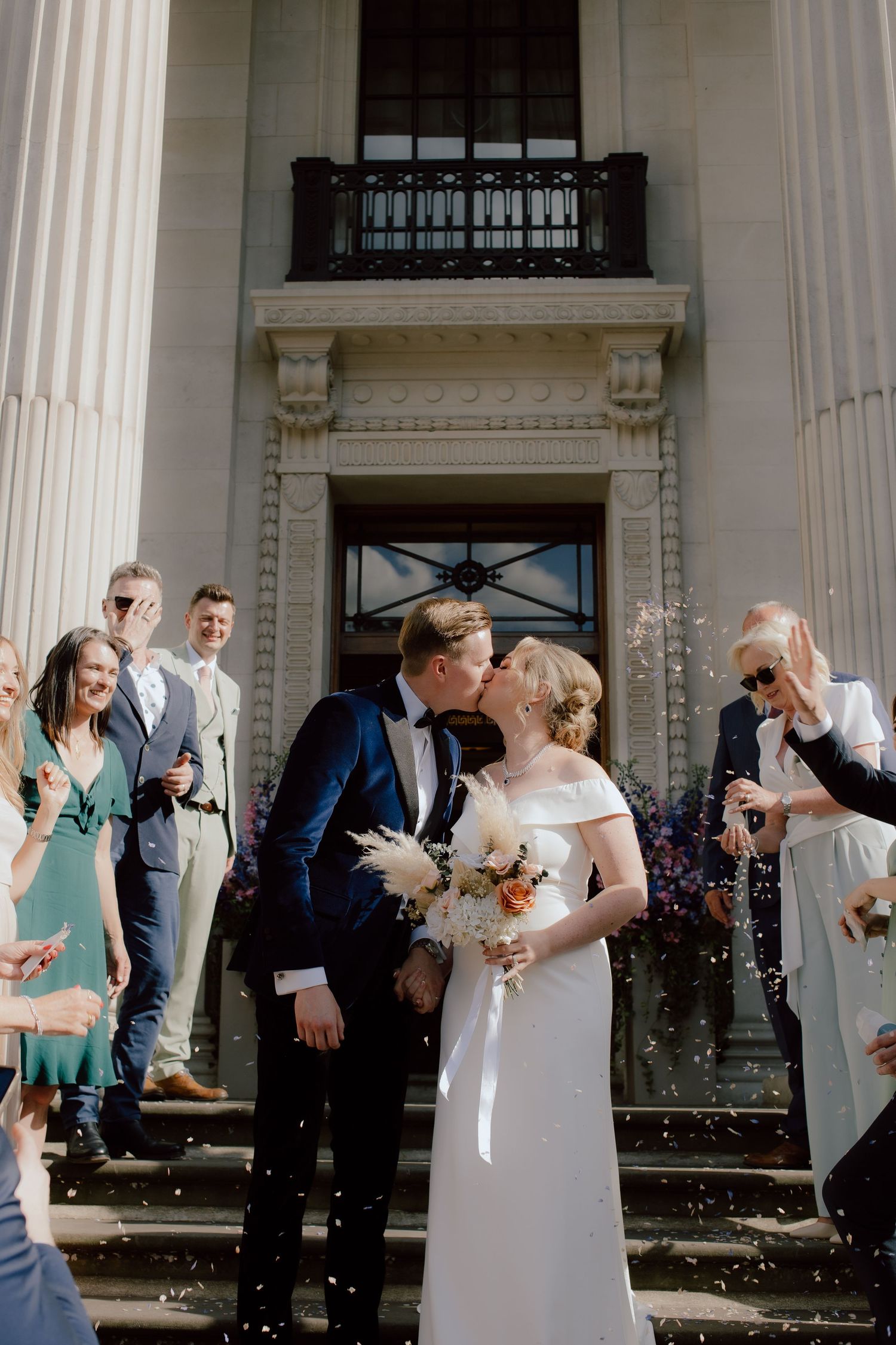 A joyful couple shares their first kiss on historic building steps while guests shower them with confetti on their wedding day.