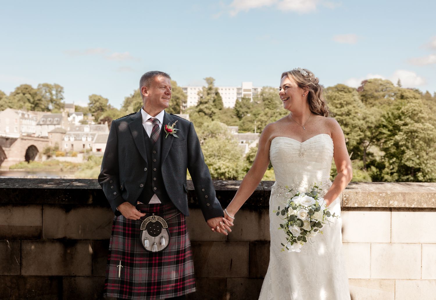 A couple in wedding attire pose together on a stone balcony with greenery and a bridge in the background.
