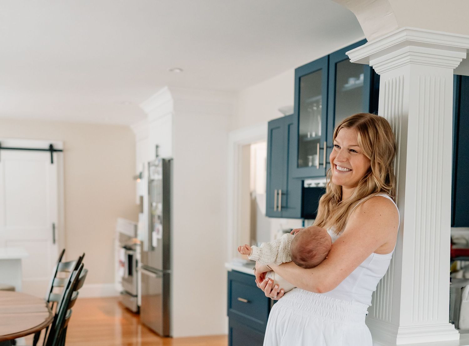 Someone in a white sleeveless dress stands smiling in a bright modern kitchen with navy blue cabinets.
