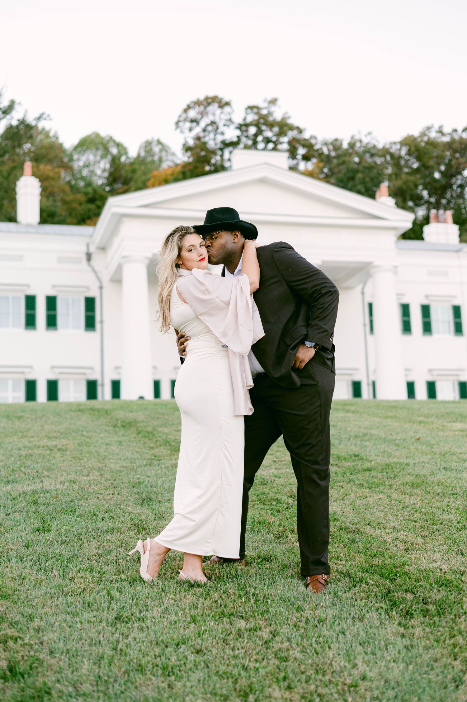 A stylish couple embracing on the lawn in front of a classic white colonial-style building.