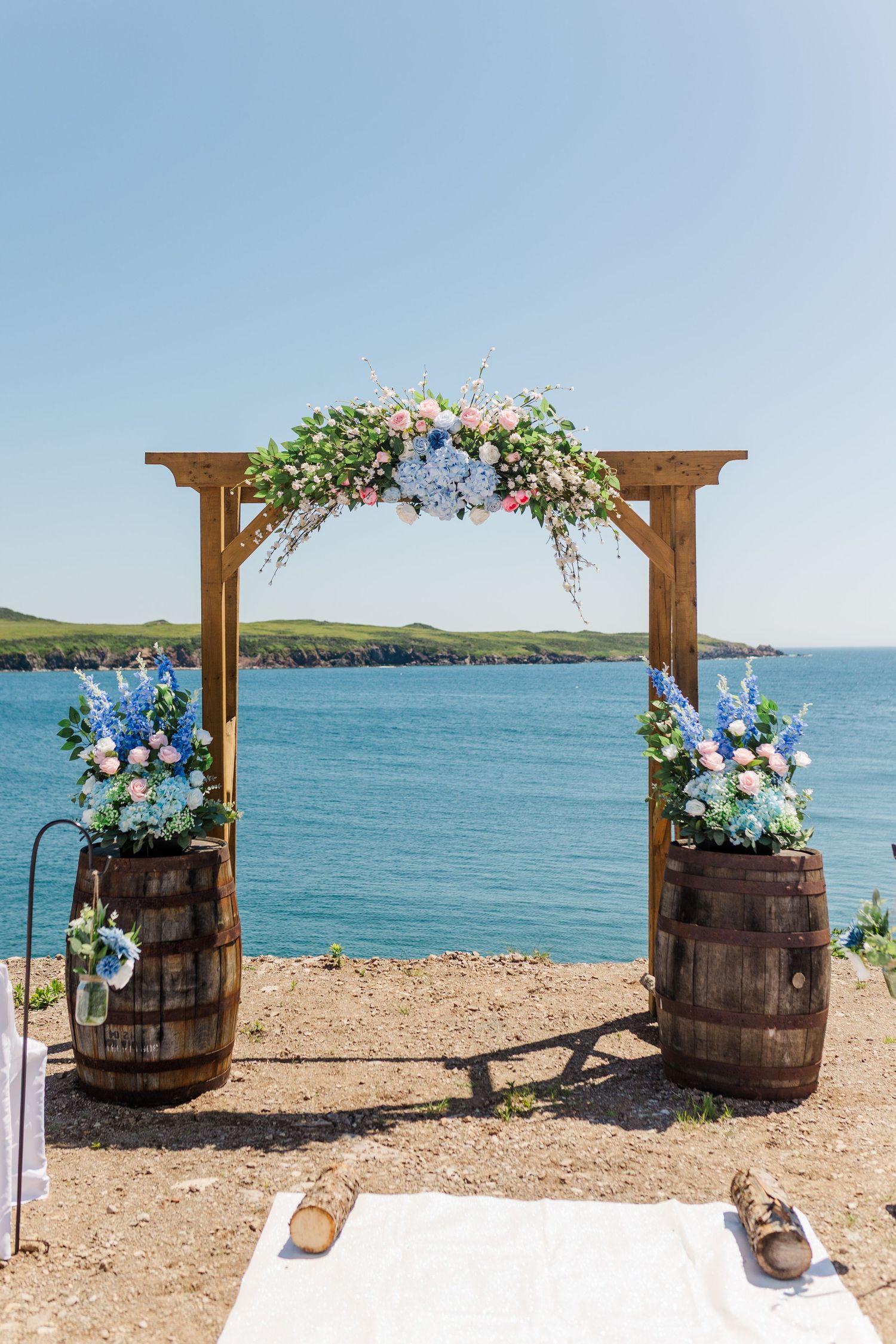 Wooden wedding arch decorated with blue and pink flowers and rustic barrels on a beach overlooking turquoise waters.