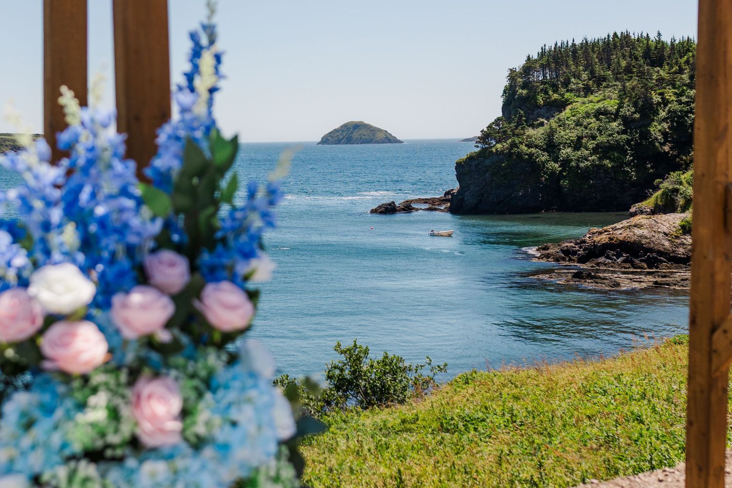 Beautiful coastal ocean view with pink and blue flower arrangement framing a rocky shoreline and small island in the distance.