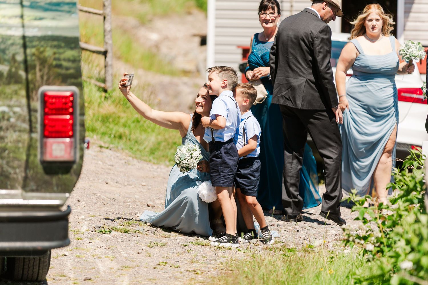 A group gathers for an outdoor summer celebration wearing formal attire, with children sitting on rustic steps.