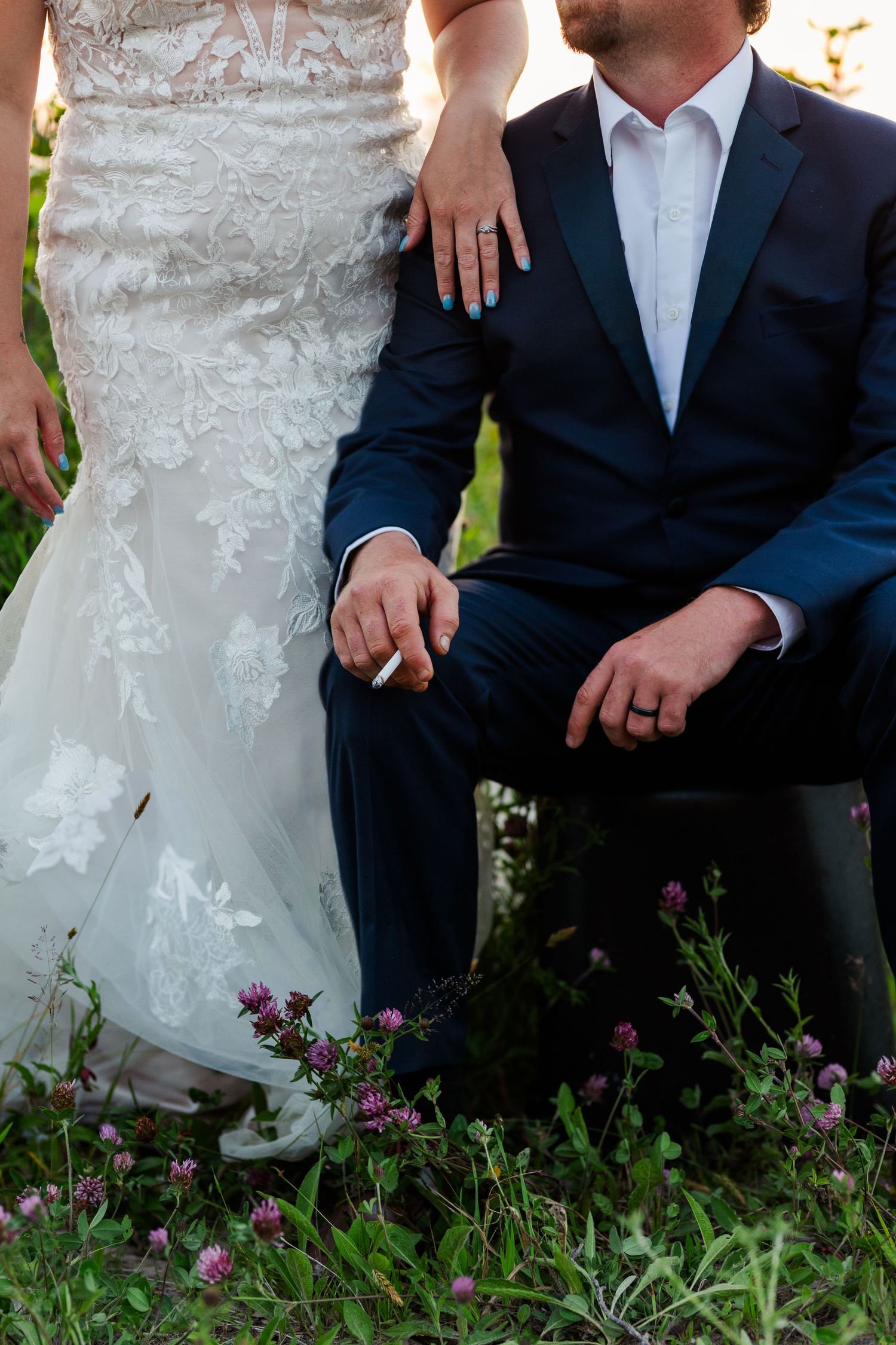 A detail shot of wedding attire with navy suits and a white lace dress amid purple wildflowers and grass outdoors.