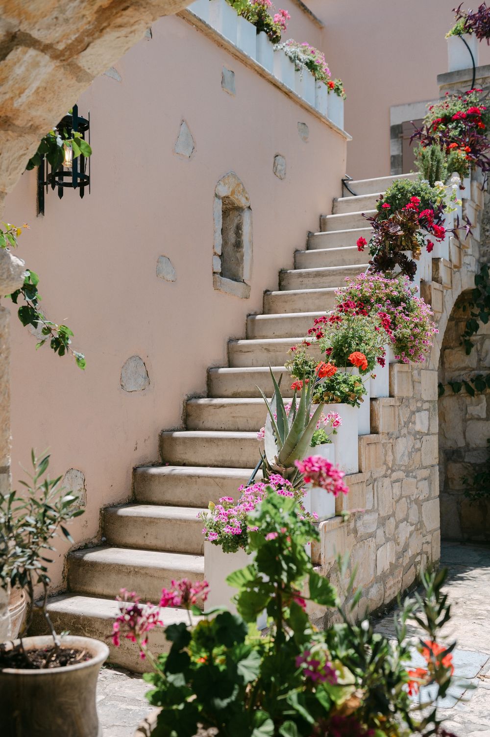 Stone staircase adorned with colorful potted flowers and climbing vines against a pink Mediterranean-style wall.
