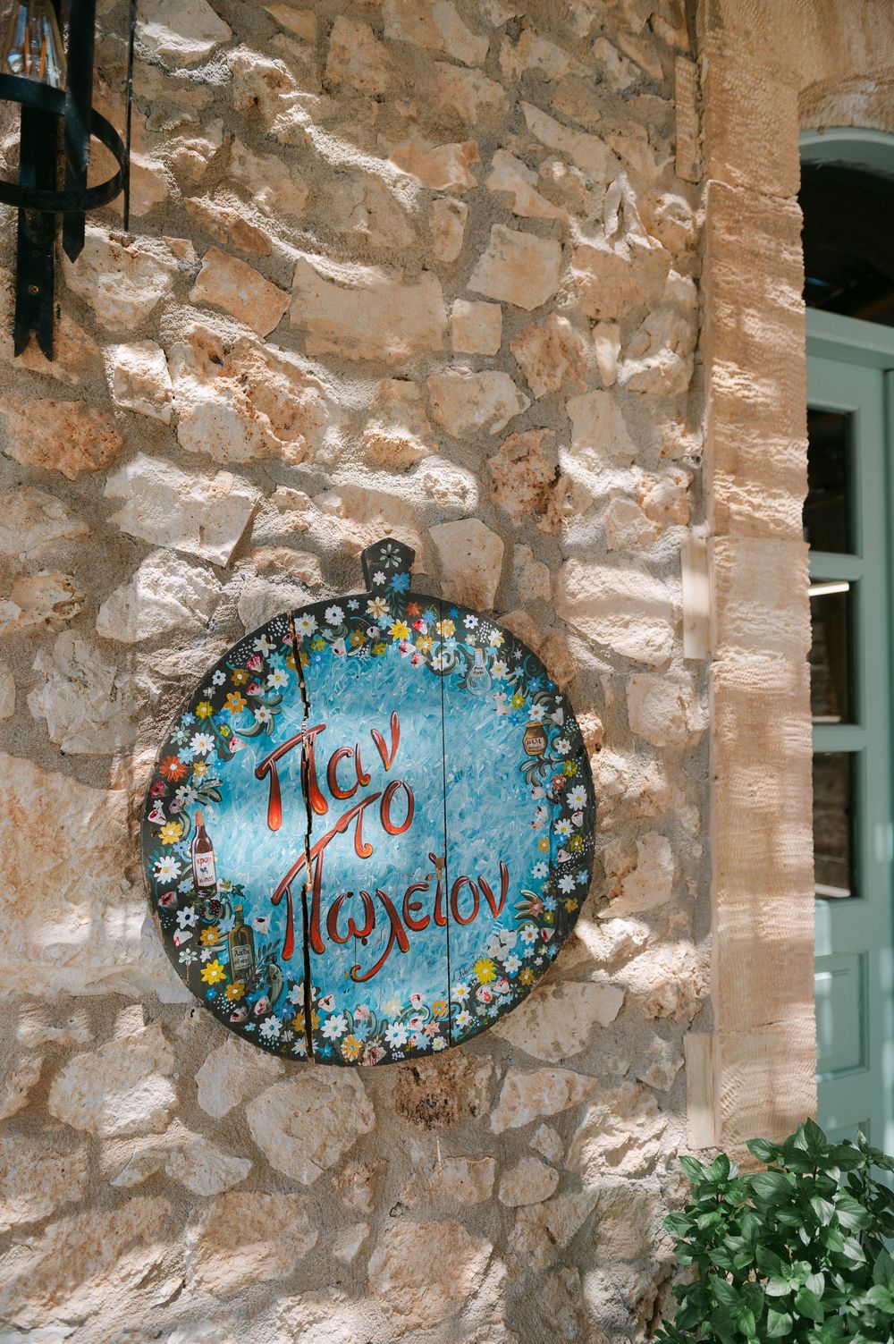 Three decorative blue and floral mosaic signs with cursive text mounted on a rustic stone wall next to green foliage.