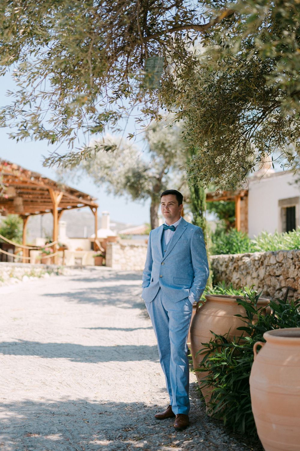 A blue-suited figure stands in a Mediterranean garden setting with terracotta pots and stone walls.