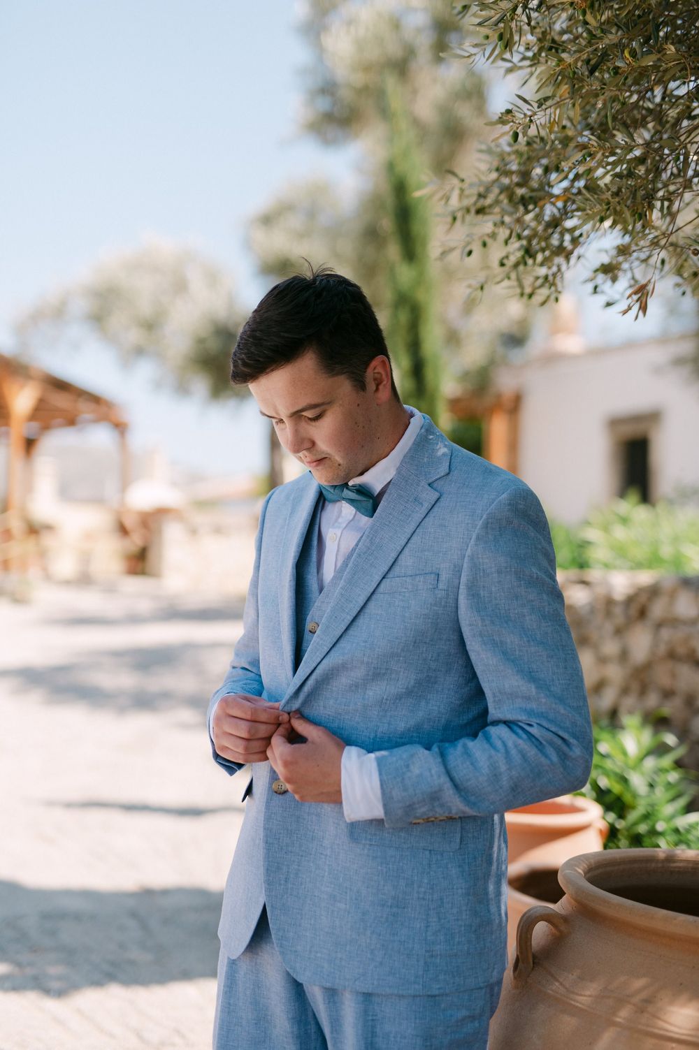 Someone in a light blue suit adjusts their jacket button while standing outside on a sunny day.