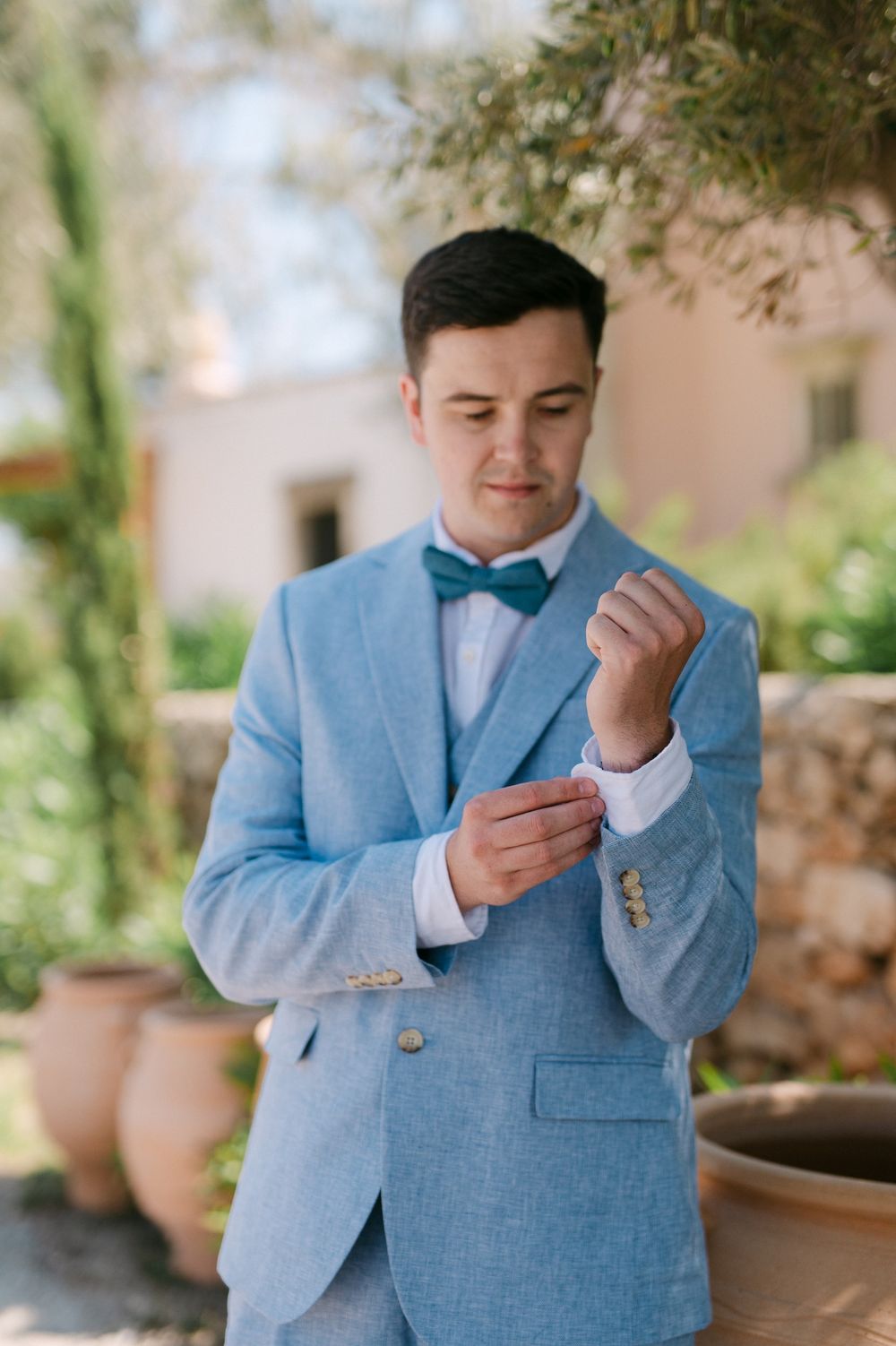 Someone adjusts their cufflinks while wearing a light blue suit in an outdoor garden setting.