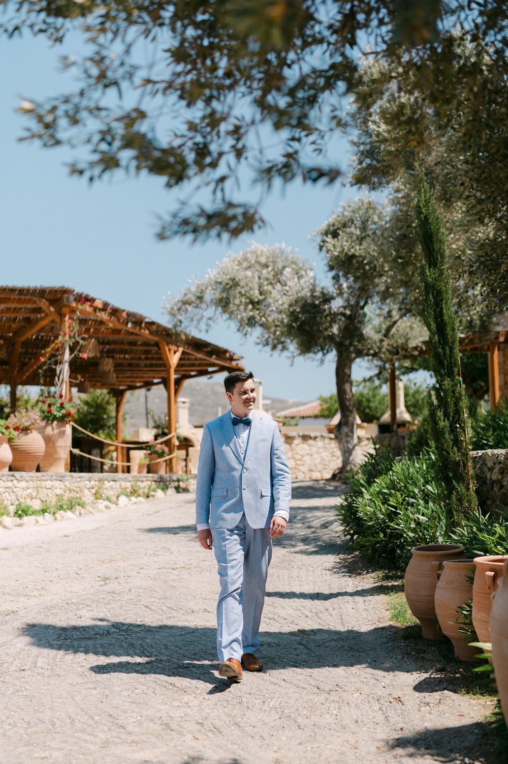 A dapper groom in a light blue suit walks along a Mediterranean garden path under a rustic pergola and olive trees.