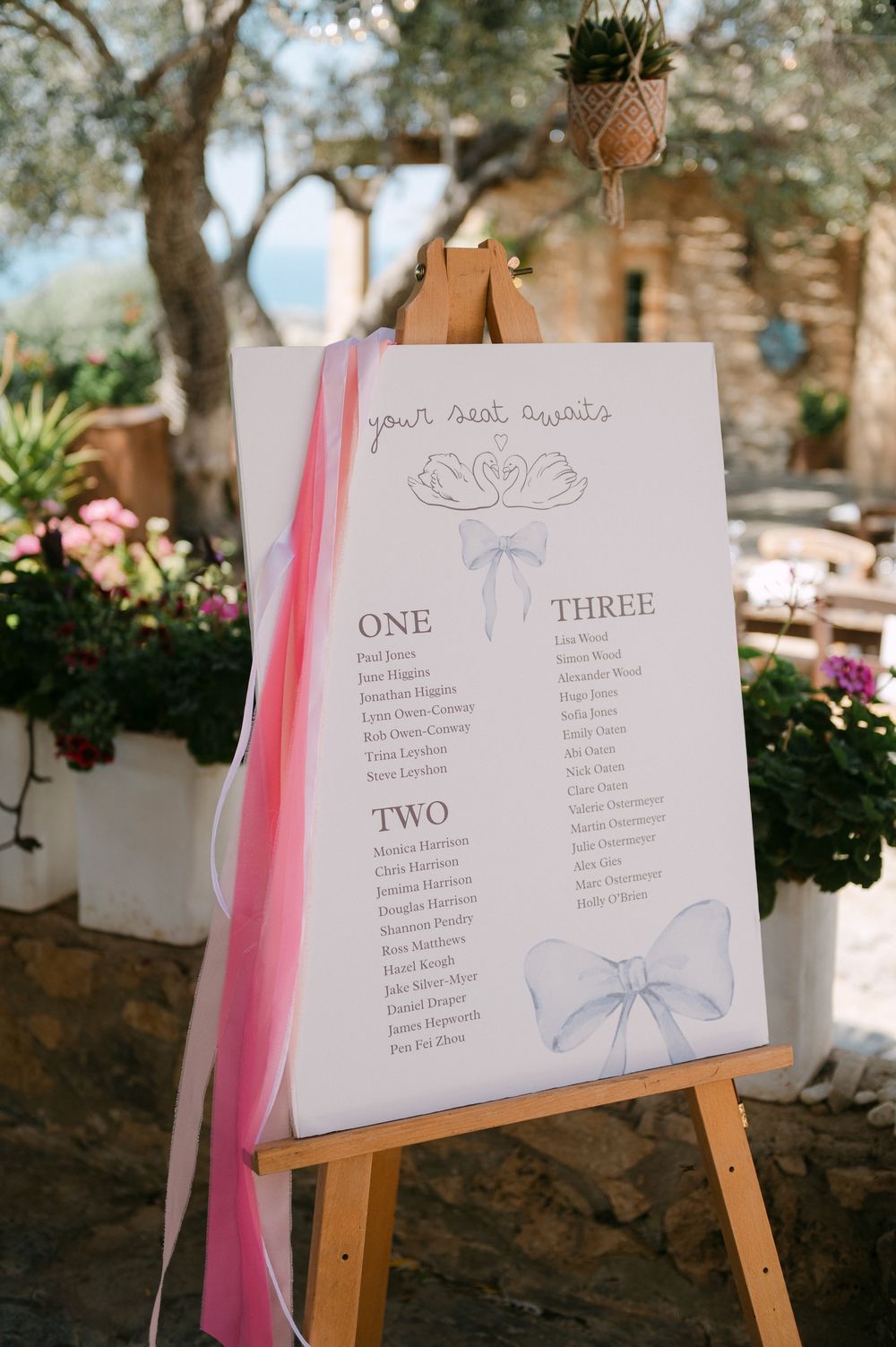 A wedding seating chart on a wooden easel with pink ribbon and blue bow decorations, displayed at an outdoor garden venue.