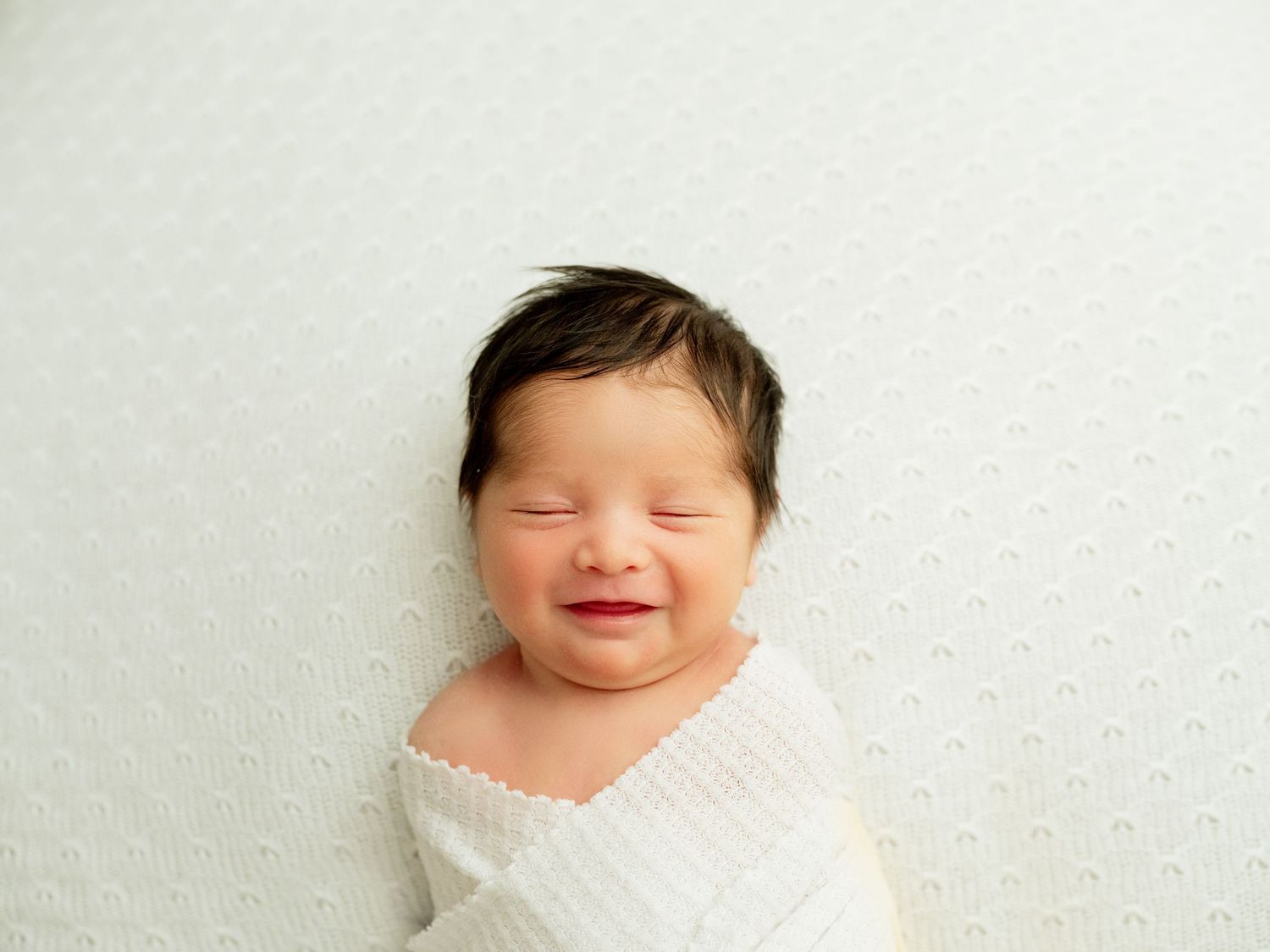 Happy newborn baby lying down with closed eyes against a plain backdrop, big milk-drunk smile.
