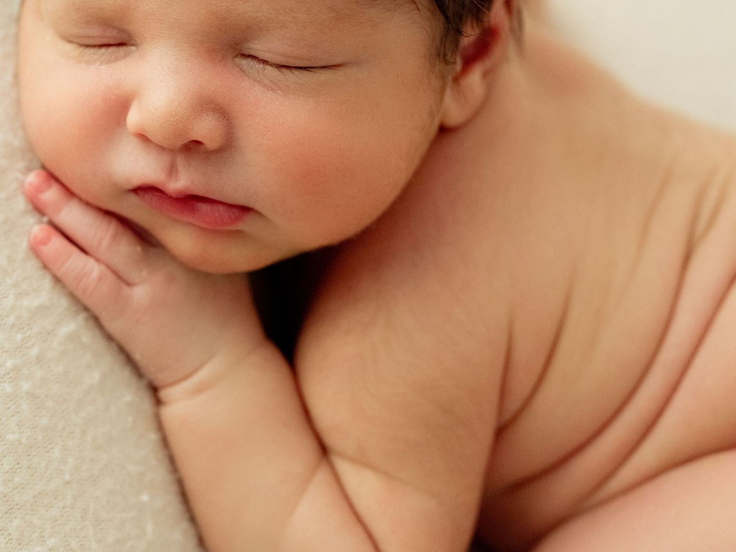 A sleeping newborn baby rests peacefully on white bedding with gentle light highlighting their delicate features. Eyelashes, cheeks and roll