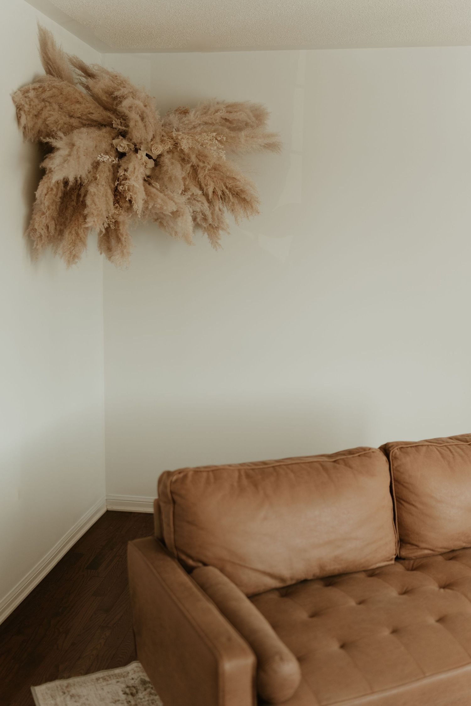 Minimalist corner featuring a tan tufted couch and dried pampas grass decor against white walls.