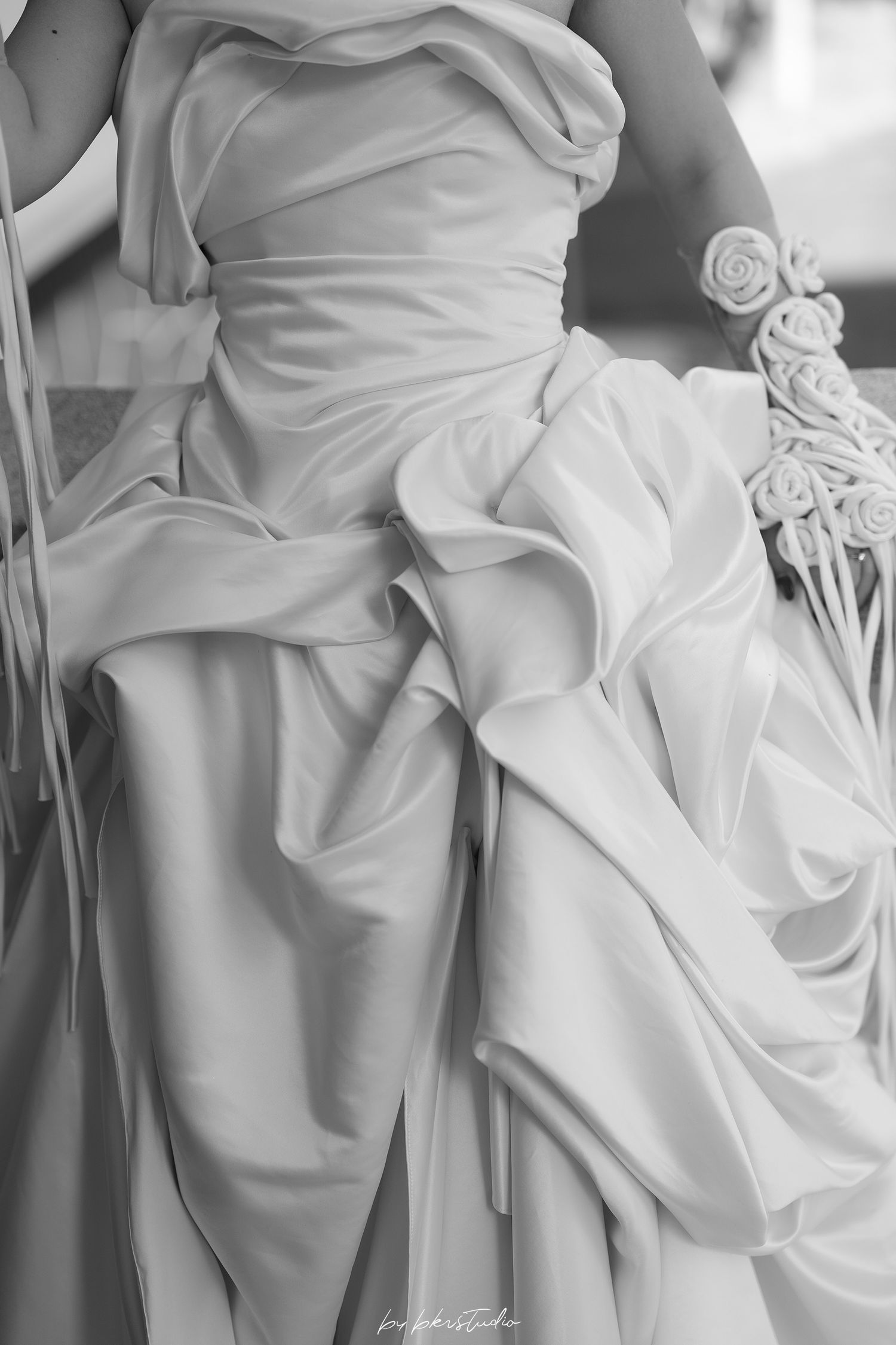 Close-up black and white photo of elegant wedding dress details showing gathered white fabric and delicate rose decorations.