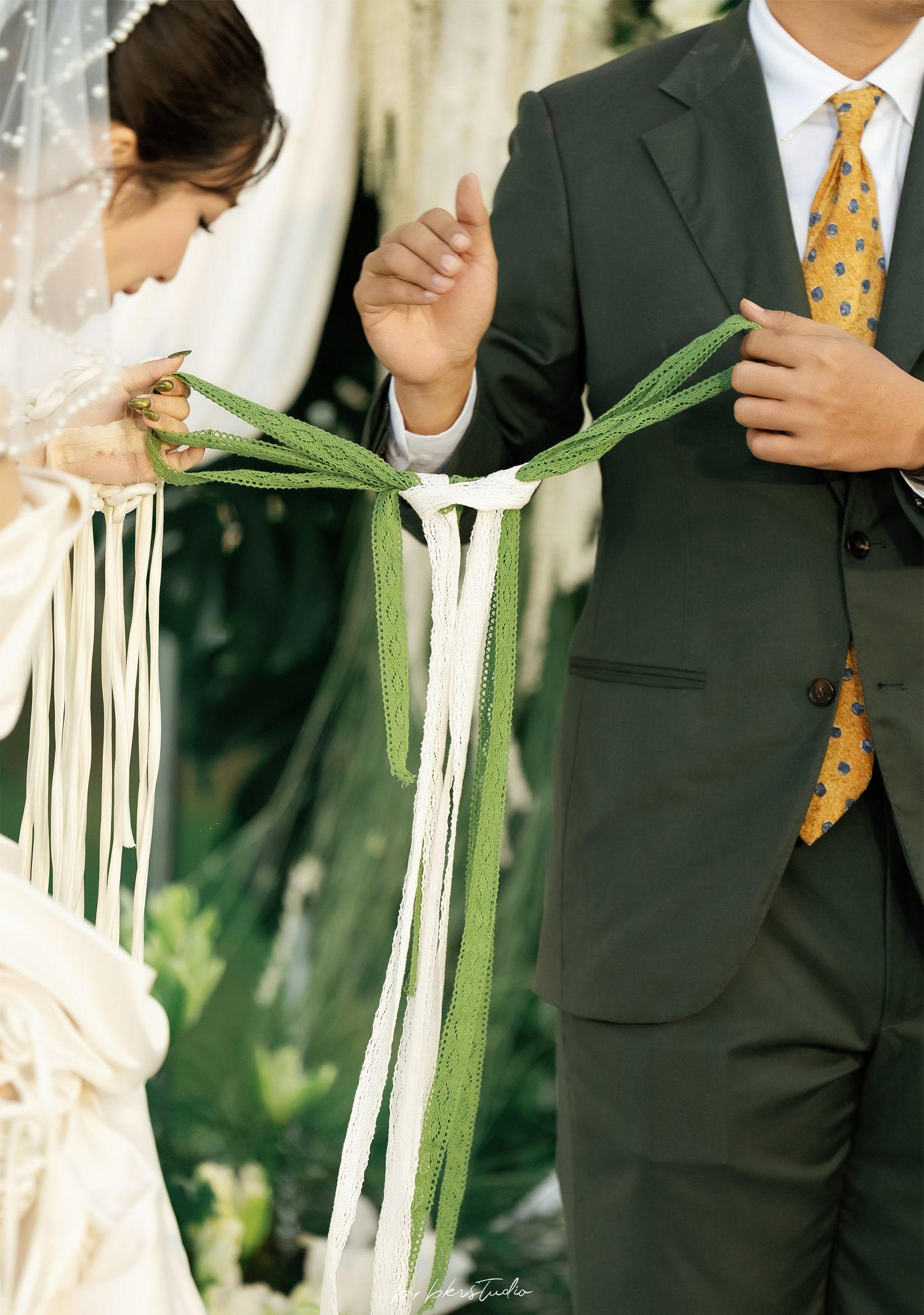 Two people in wedding attire hold white and green ribbons during a ceremonial handfasting ritual outdoors.