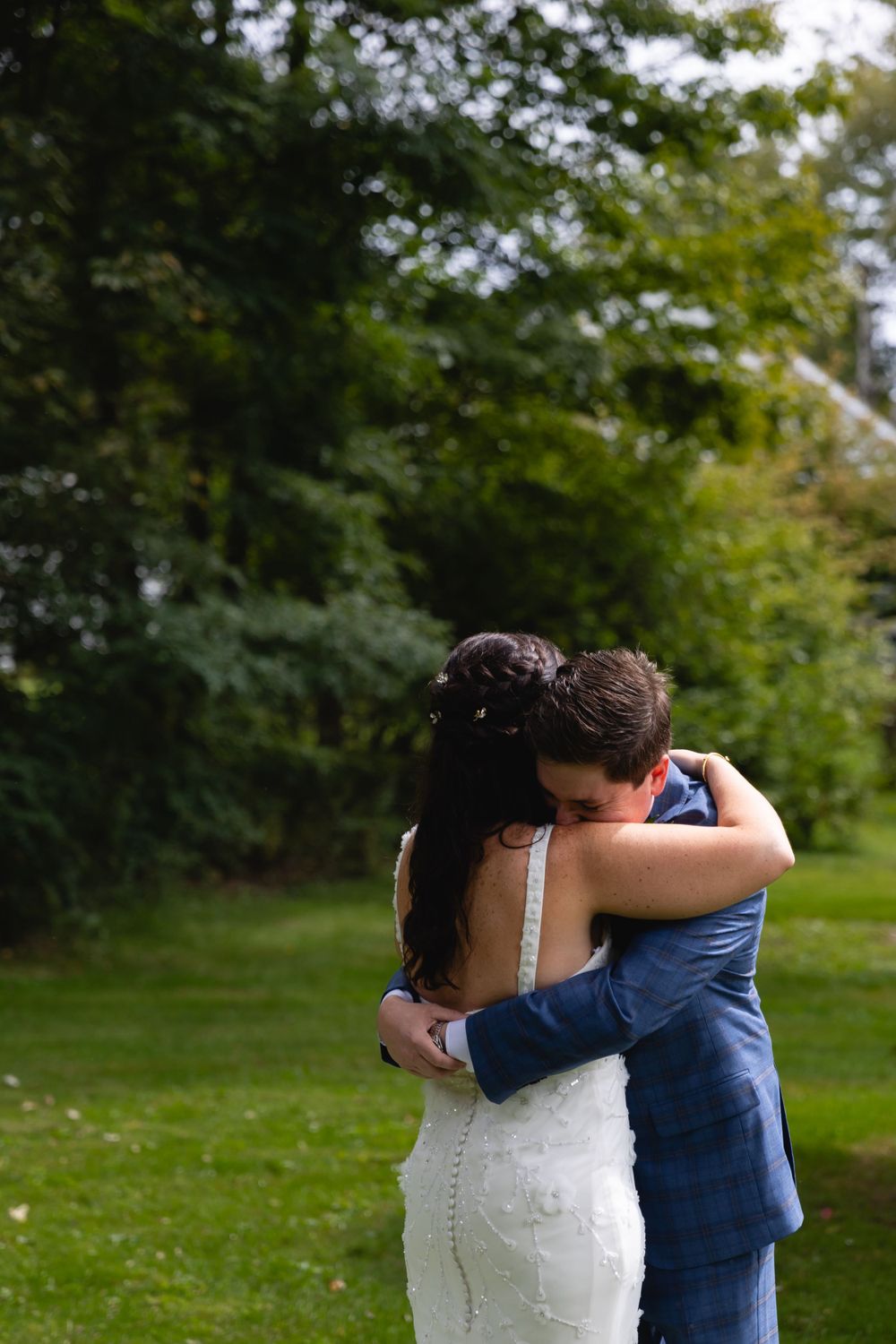 A couple in wedding attire share an intimate embrace on a lush green lawn surrounded by trees.