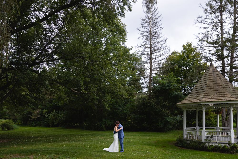 A couple embraces near a white gazebo surrounded by lush green trees and grass in a scenic outdoor setting.