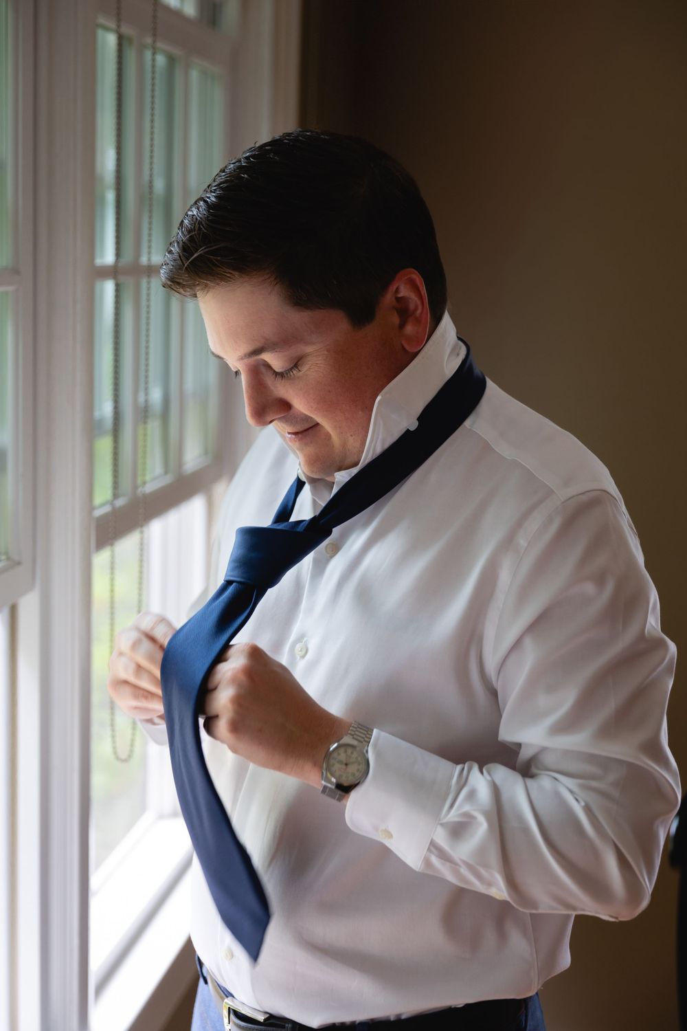 Someone adjusts their navy blue necktie while wearing a crisp white dress shirt by a window in natural light.