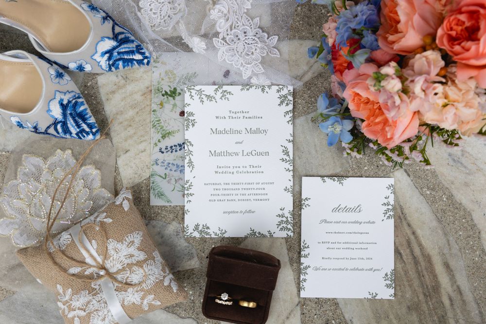 Wedding details including invitations, gold rings in black box, traditional blue and white shoes, and coral roses on lace.
