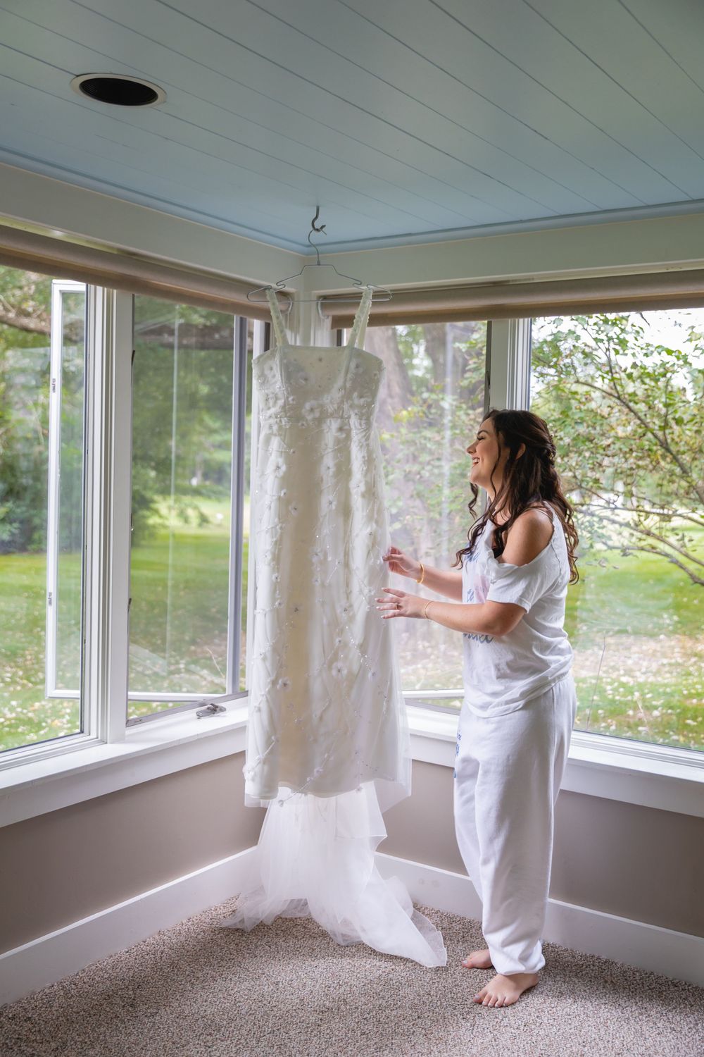 A woman in white clothing admires a lace wedding dress hanging in a sunlit room with large windows and natural light.