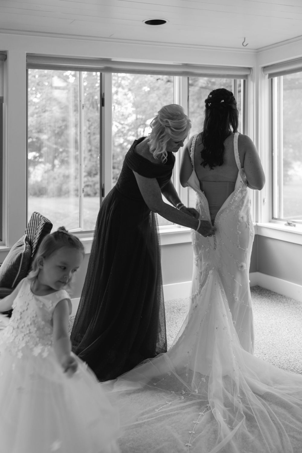A black and white photo capturing a quiet moment of wedding dress preparation in a sunlit room.