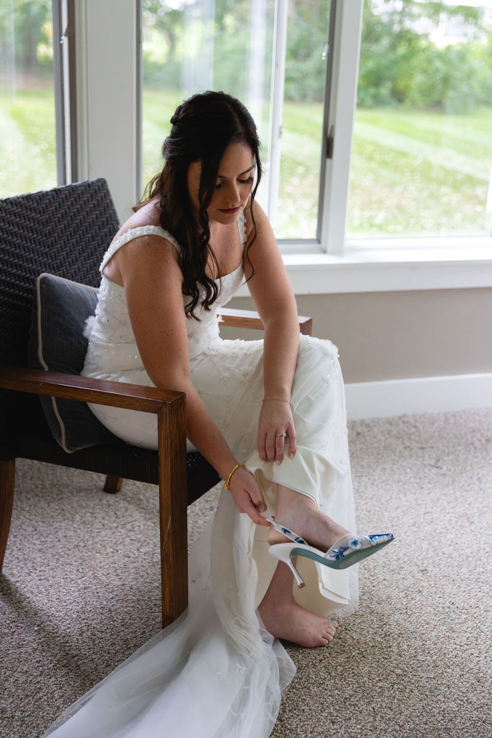 A bride in a white wedding dress sits in a wooden chair by a window, putting on her blue shoes.