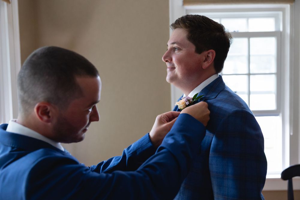 A person helps adjust a bright blue plaid suit jacket and tie in a naturally lit room by a window.