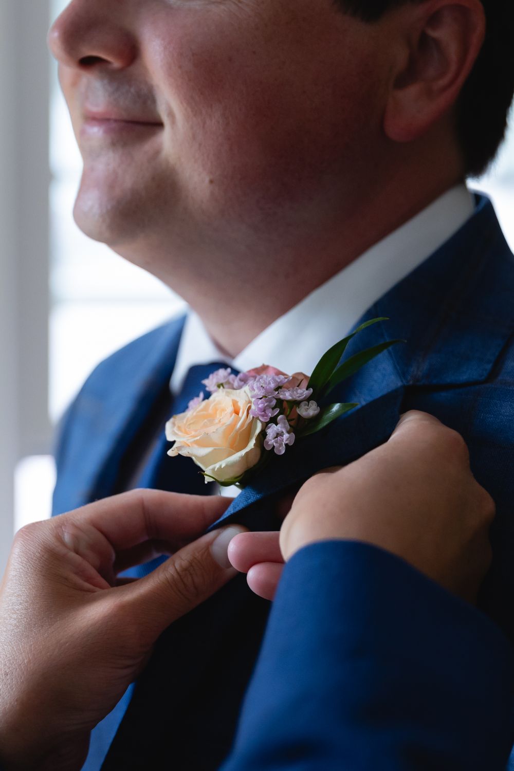 Someone wearing a navy blue suit gets a peach rose and small pink flower boutonniere pinned to their lapel.