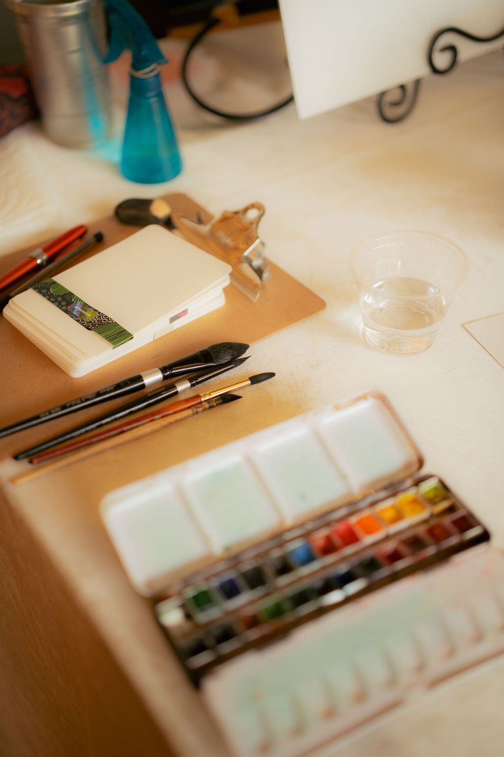 Artist workspace with watercolor palette, brushes, clipboard and art supplies on a beige surface.