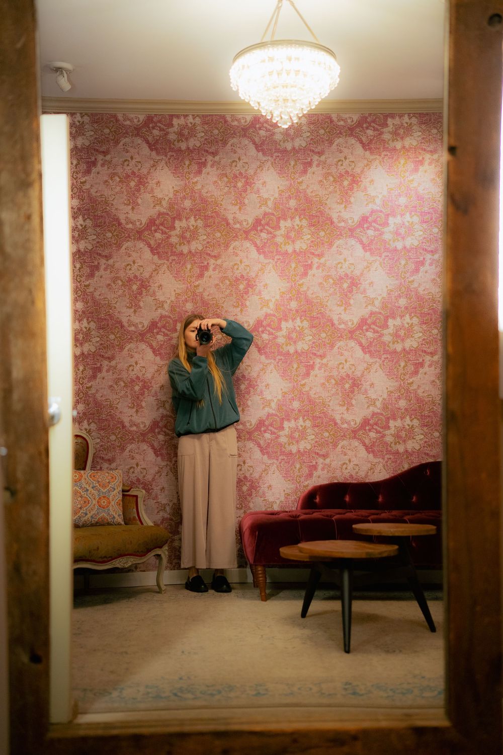 A room with pink damask wallpaper features a crystal chandelier, red velvet seating, and someone taking a photograph.