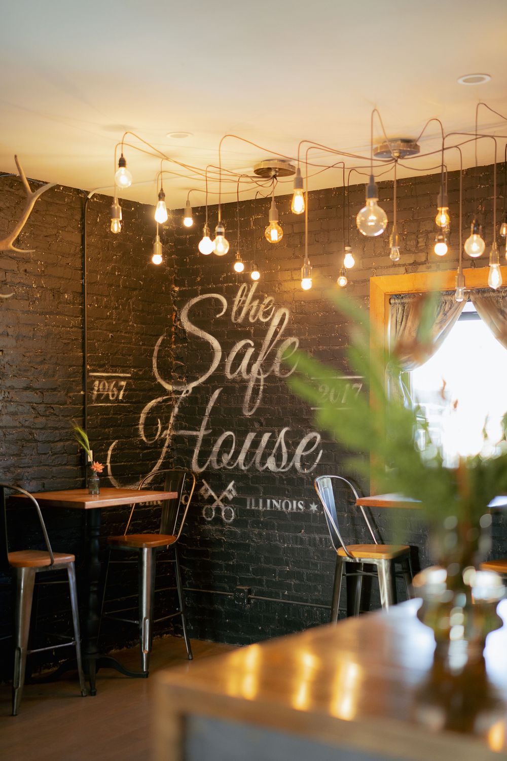 Industrial cafe interior with hanging Edison bulbs and 'The Safe House' written in script on a dark painted brick wall.