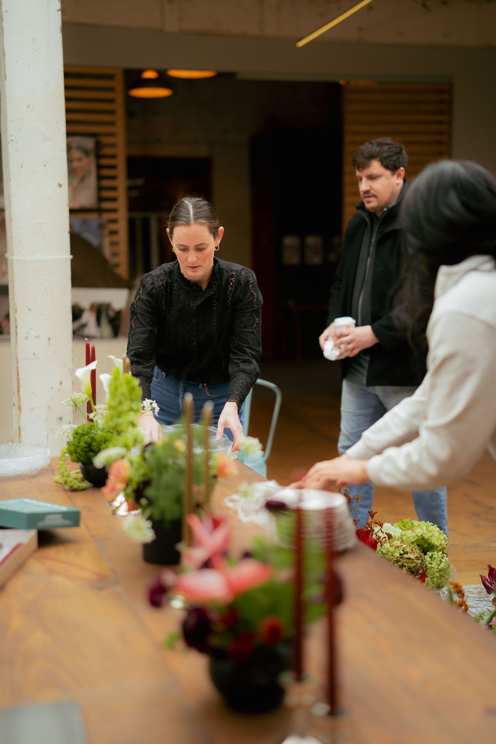 People gather around a wooden table with plants and floral arrangements in an indoor setting.